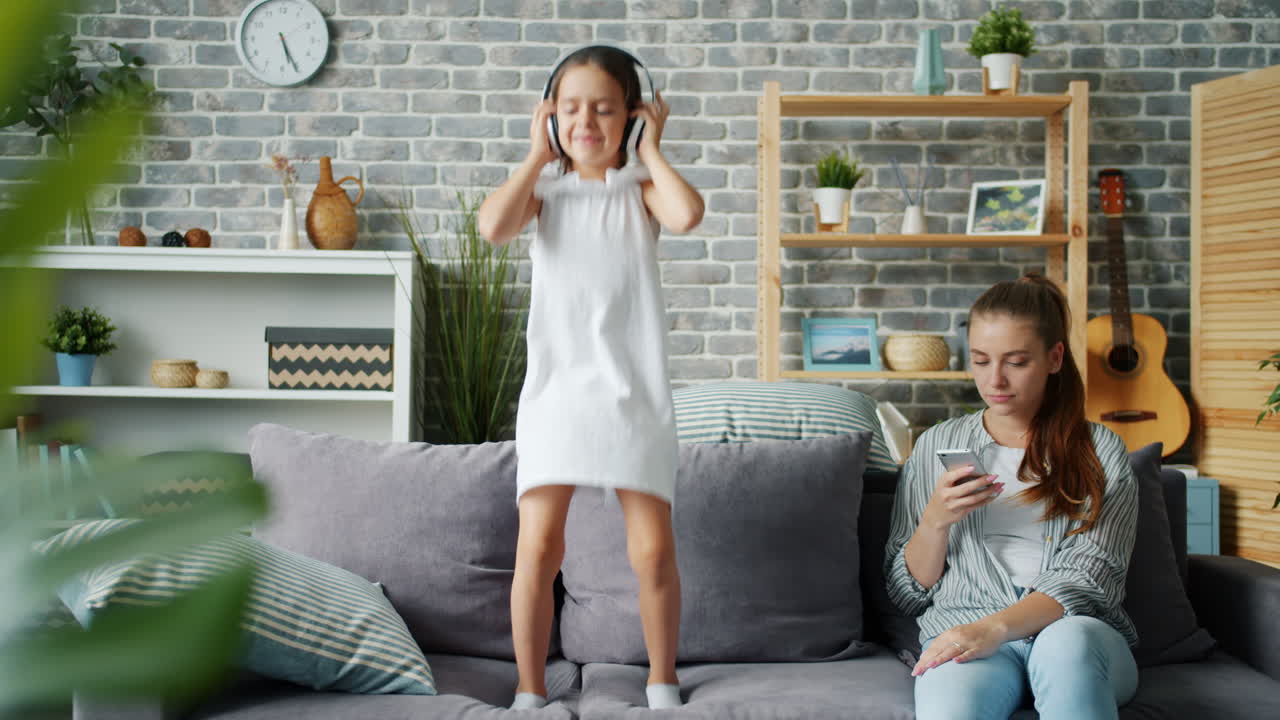 Daughter Dancing with Headphones on Sofa while Mom Uses Phone
