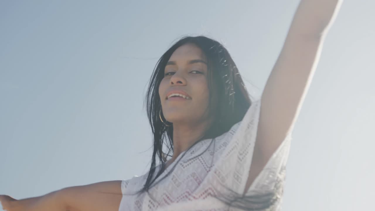 Happy hispanic woman dancing with arms outstretched on sunny beach, smiling, slow motion
