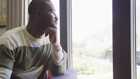 video of thoughtful african american man sitting and looking out of window at home