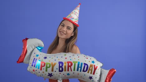 una mujer joven posa con un letrero de globo de feliz cumpleaños y un sombrero de fiesta, aislada en un fondo de estudio azul