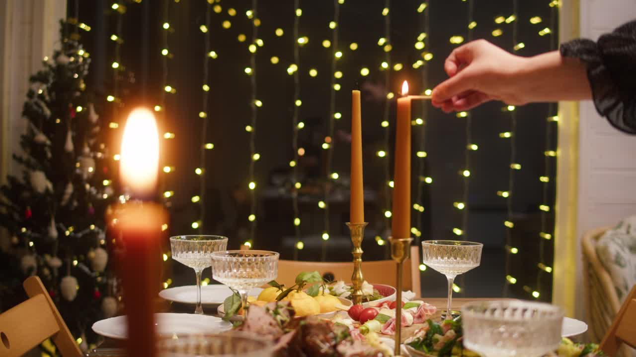 Woman lighting candles on dining table close-up. Friends celebrating new year, delicious dishes, cooked homemade food on plates. Christmas party, festive dinner at home.