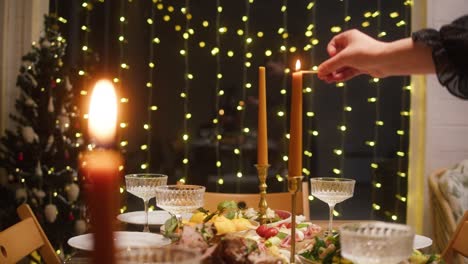 woman lighting candles on dining table close-up. friends celebrating new year, delicious dishes, cooked homemade food on plates. christmas party, festive dinner at home.
