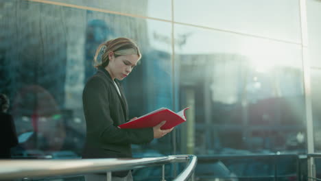 woman in black suit reads red-covered book thoughtfully near glass building with sunlight ambiance reflecting across glass panels, creating serene, reflective mood in urban setting
