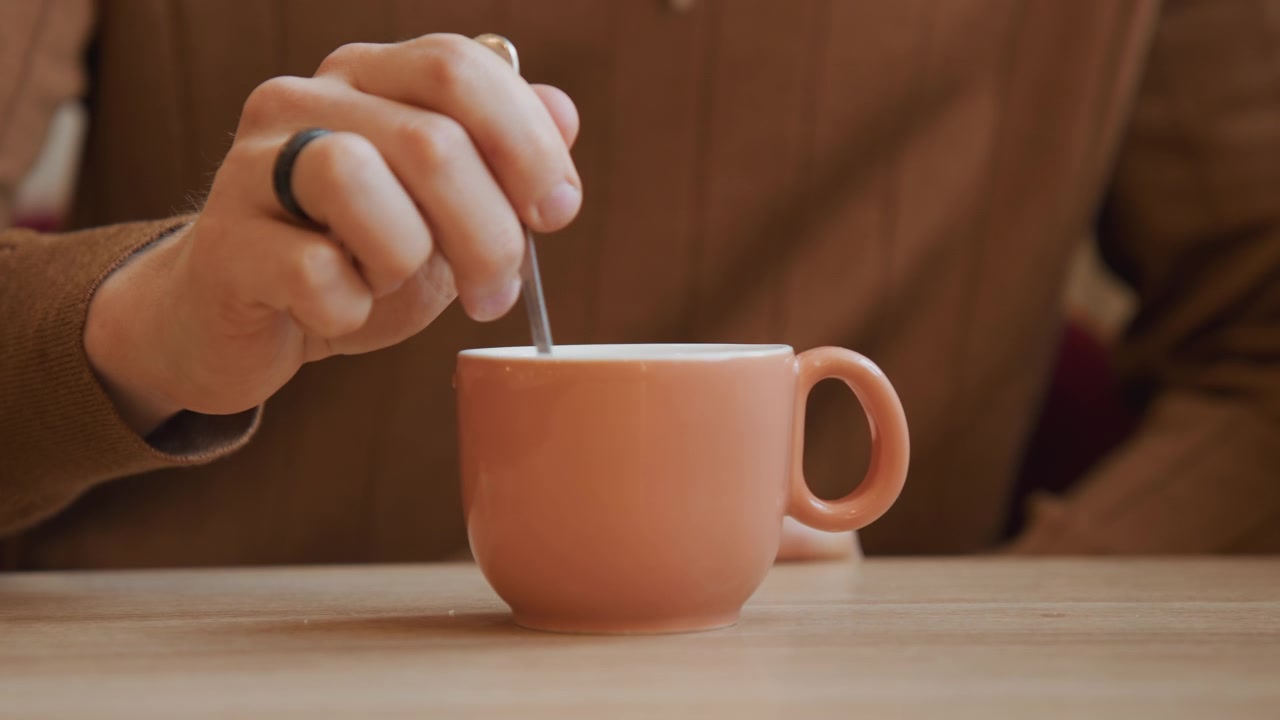 Close up of hand stirring liquid in pink ceramic cup on wooden table. person wearing brown sweater and ring repeatedly stirs drink with spoon throughout sequence. Soft lighting creates warm