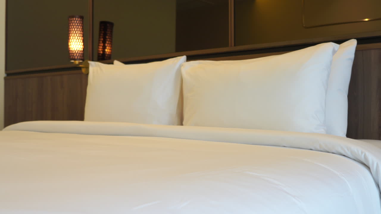left to right panning shot of a very clean and comfortable hotel room bed with a pure white fluffy comforter and soft pillows against a dark wood panel wall.
