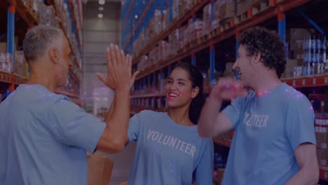 Animation-of-glowing-spots-of-light-over-diverse-people-high-fiving-volunteering-in-warehouse,