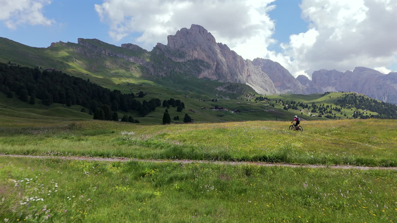 En cyklist cyklar genom livfulla ängar med en reflekterande sjö och höga bergstoppar i Dolomiterna, Italien, en ljus dag