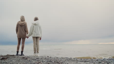 mother and daughter look at the ocean, where storm clouds are in the distance. standing on the shore, holding hands