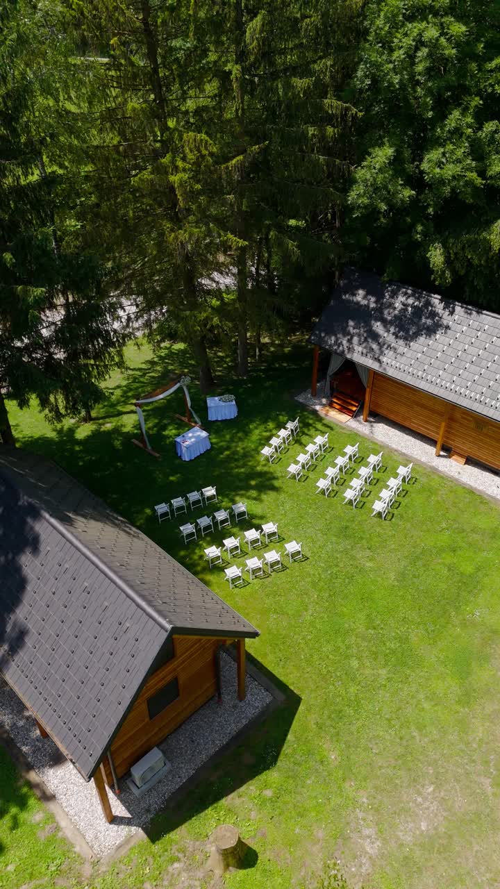Vertical, aerial: wedding ceremony with an arch and white chairs during the day in the forest, orbit drone shot