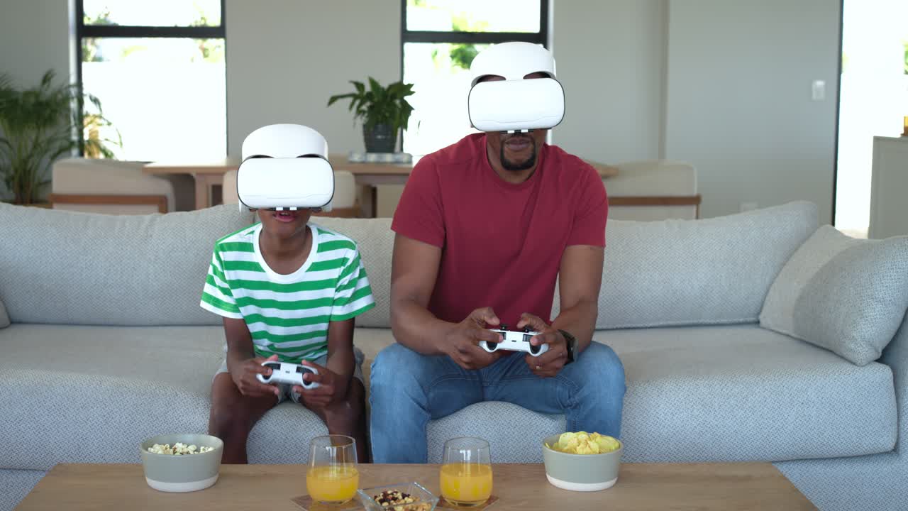 Padre e figlio afroamericani che indossano visori VR bianchi sul divano dopo l'inizio di una partita con i controller