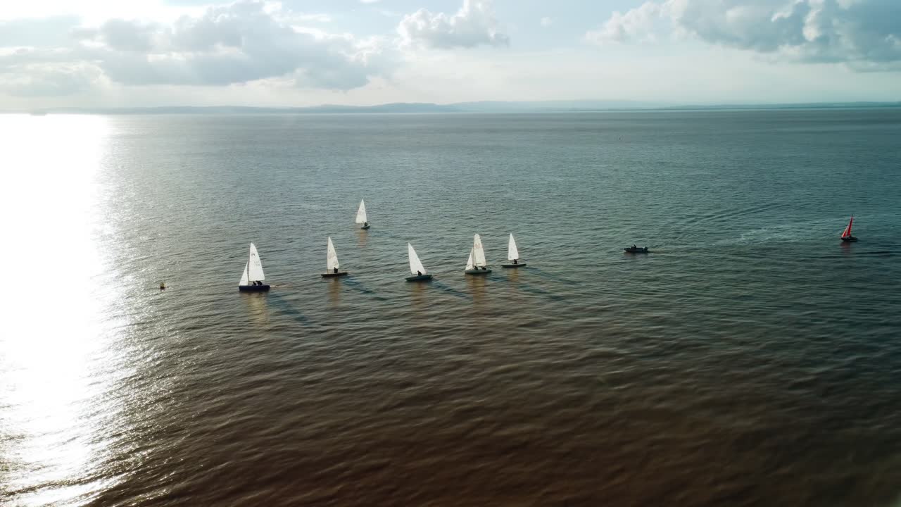 Ripresa con drone di piccole barche a vela al largo della costa di Clevedon, Regno Unito, con il sole che tramonta dietro di esse. Mentre le barche superano una boa, il drone esegue una panoramica a sinistra per rivelare il molo di Clevedon e la costa