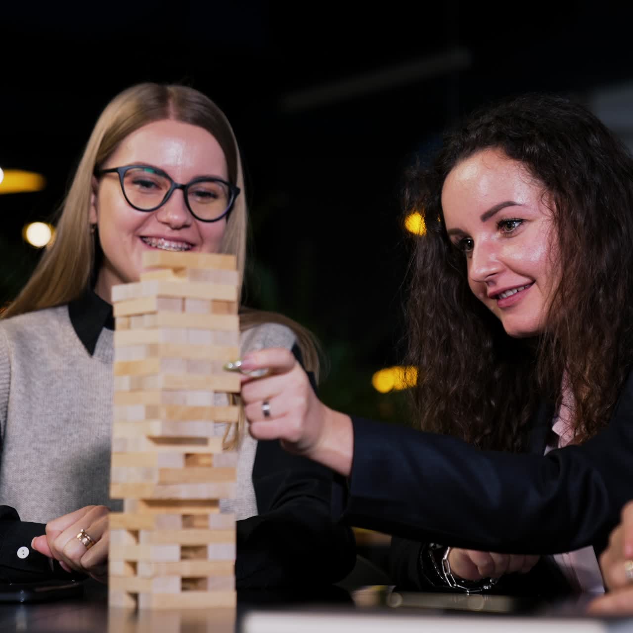 Una giovane e bellissima donna dai capelli lunghi estrae un mattone di legno da una pila. Una signora bionda seduta accanto la guarda sorridendo con attenzione