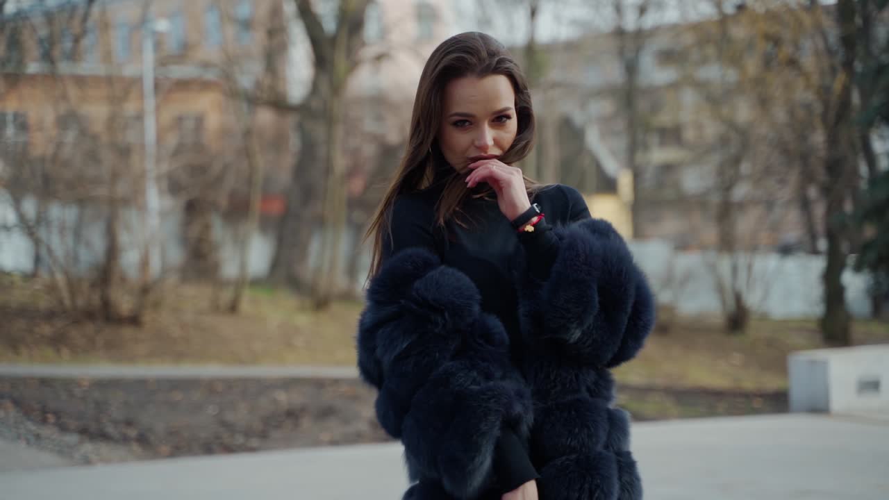 Young beautiful fashionable lady wearing stylish dark fur coat posing in park. Model looking aside. Female fashion concept. City lifestyle. Fashion video.