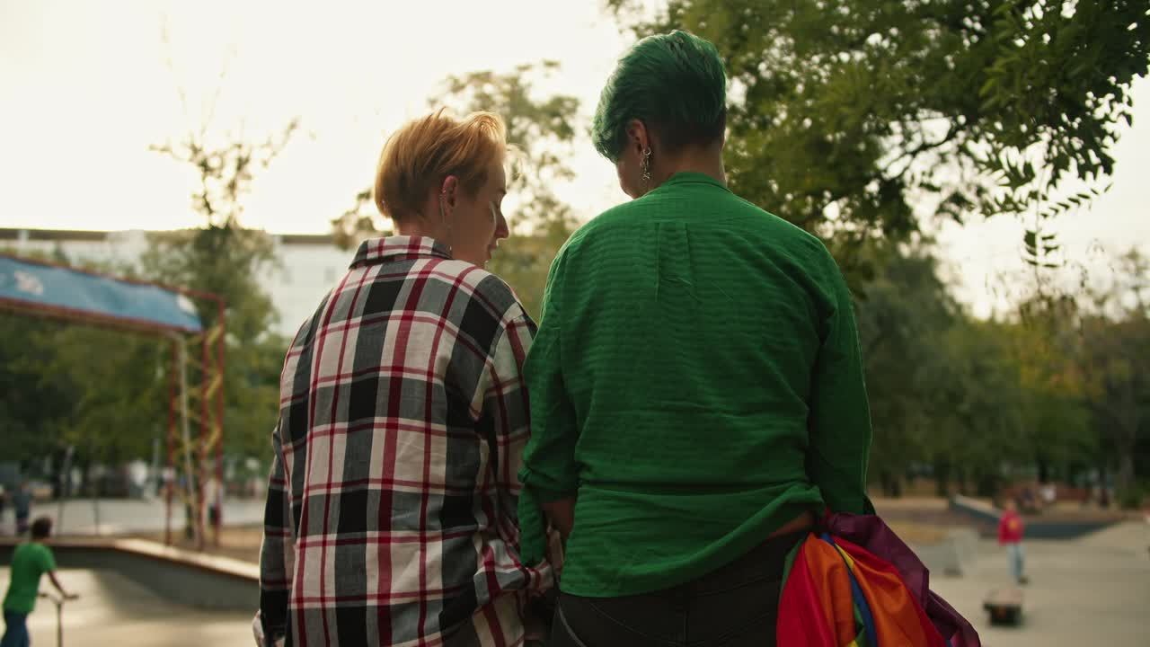 Rear view of a girl with green hair and a short haircut in a Green shirt and a blonde girl with a short haircut in a checkered shirt are sitting on a fence in a skate park on their date and talking to each other