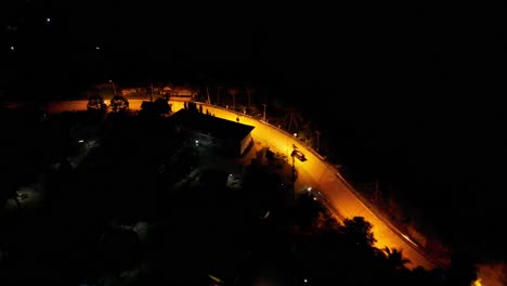 Aerial-drone-shot-of-a-car-driving-down-an-empty-road-at-night-in-a-rural-area-with-yellow-street-lights