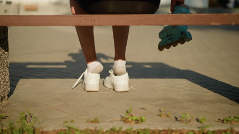 back view of lady's leg partially out of white sneakers while sitting on bench, reaching for roller skates to wear, sunlight casts soft shadows on pavement