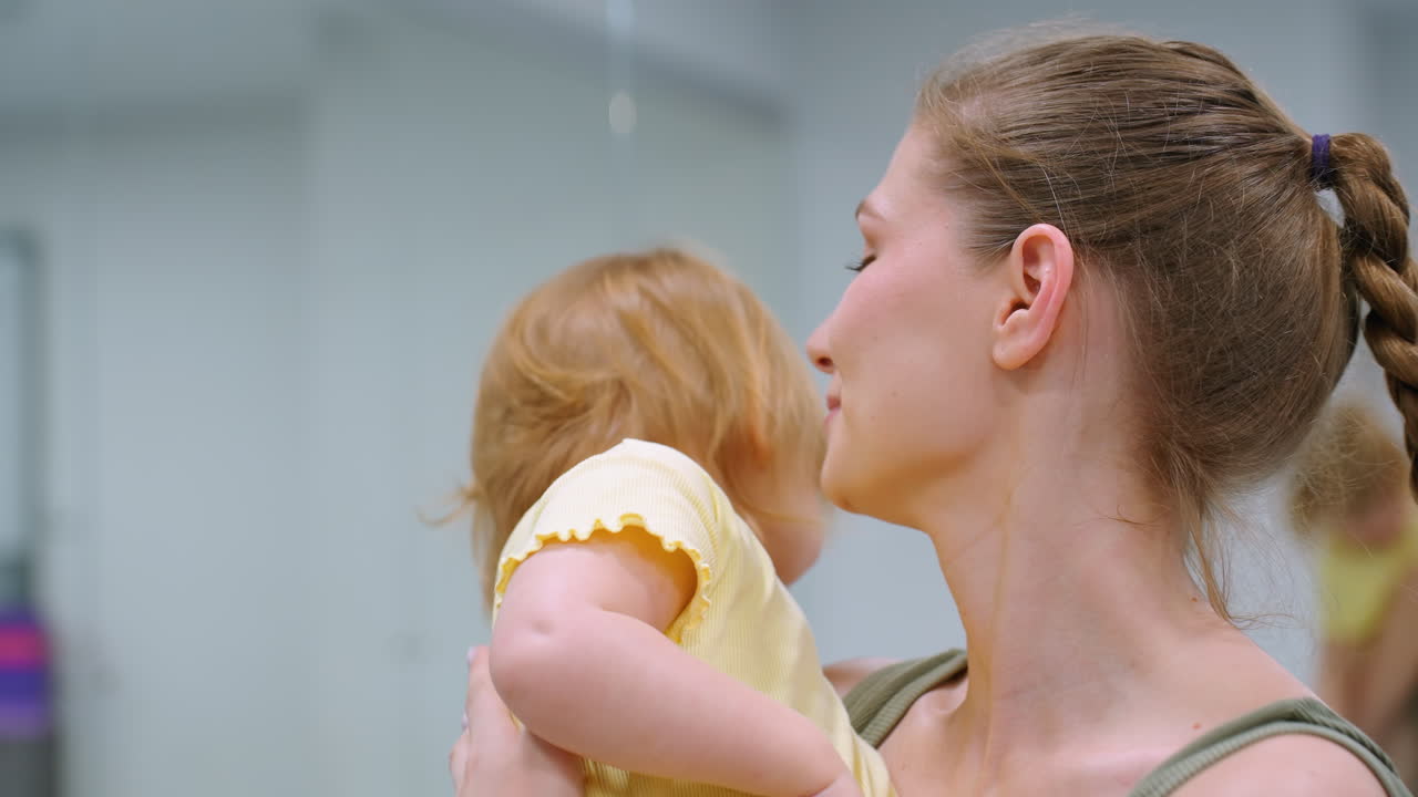 즐거운 이모와 조카가 잘 조명된 체육관에서 행복한 순간을 나누고, 아이는 뒤로 돌아서며 장난스럽게 작별 인사를 하며, 큰 거울이 그들의 등을 반영하고, 약간 흐릿합니다.