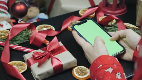 teléfono con pantalla verde en la mesa. los dedos femeninos están escribiendo mensajes en el teléfono celular con pantalla verde. pantalla chromakey. manos usando teléfono inteligente con pantalla verde y viendo regalos de navidad