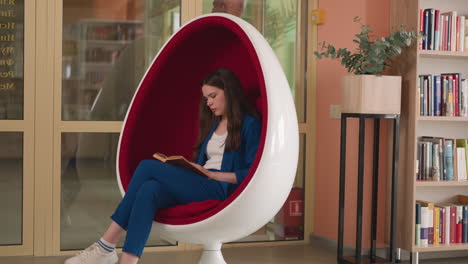 woman reads book in egg chair in library. concentrated brunette lady rests with old female novel in comfortable armchair. contemporary reading hall