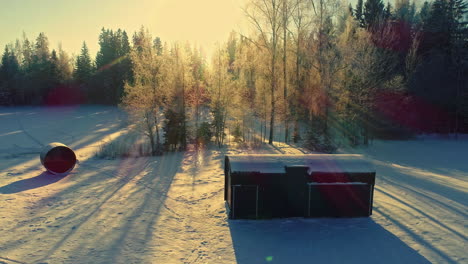 aerial view snowy field with people approaching wooden cabin, sunlight peaking through trees during snowfall