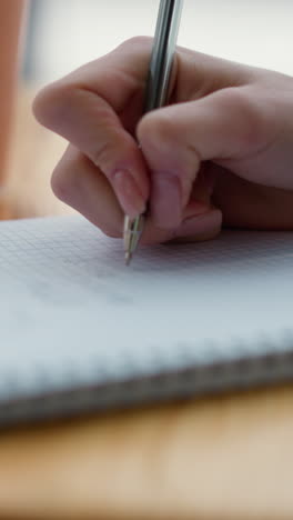 primer plano de la mano del estudiante escribiendo en papel gráfico con un bolígrafo, taza de café y teléfono borroso en el fondo, capturando el estudio, el enfoque y la atmósfera académica