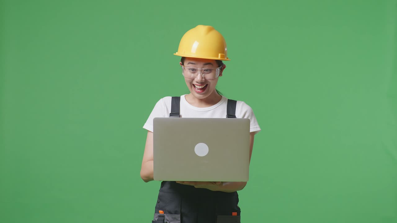 안경과 안전 헬을 착용 한 아시아 여성 노동자가 노트북을보고 녹색 화면 배경 스튜디오에 서서 축하하는 골을 비명을 지르고 있습니다.