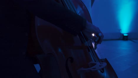 a musician dressed elegantly plays the cello in a room with blue light and a bright light in the background, in slow motion