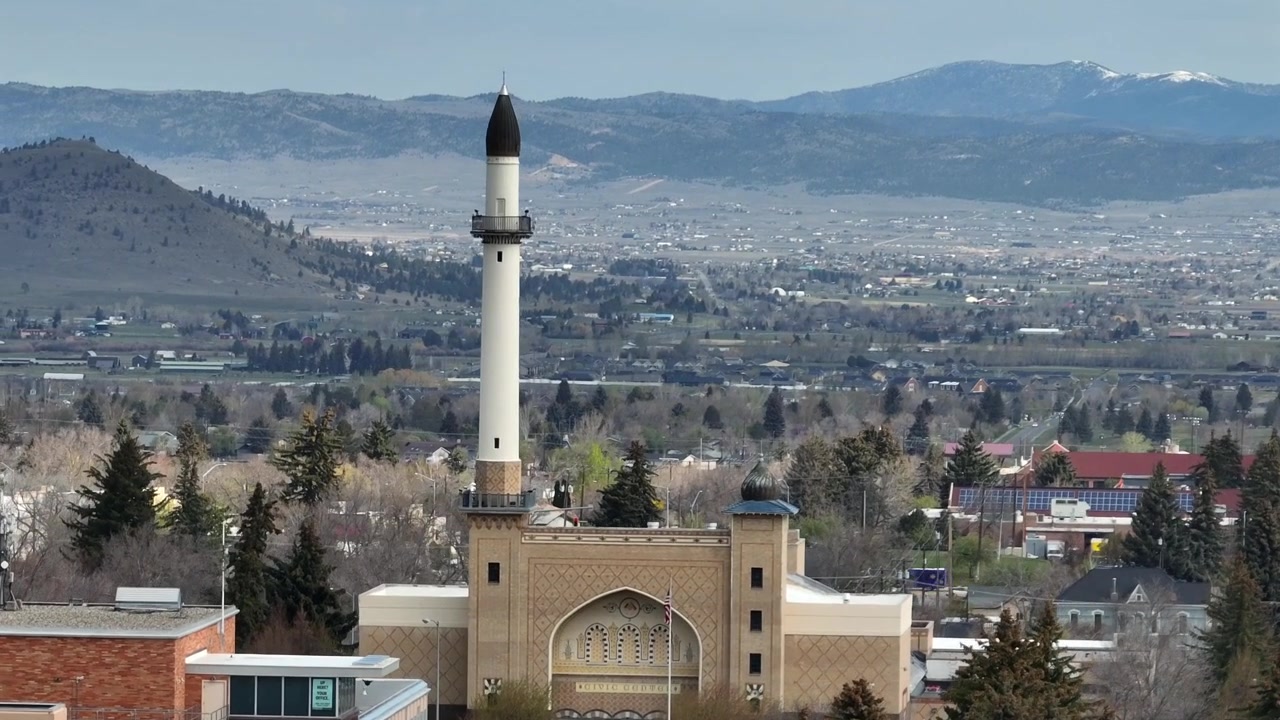 Moorish-style minaret with black dome caps tan building, horseshoe arch entrance, surrounded by evergreen trees overlooking valley and mountains. Helena, Montana, USA. Aerial drone wide shot