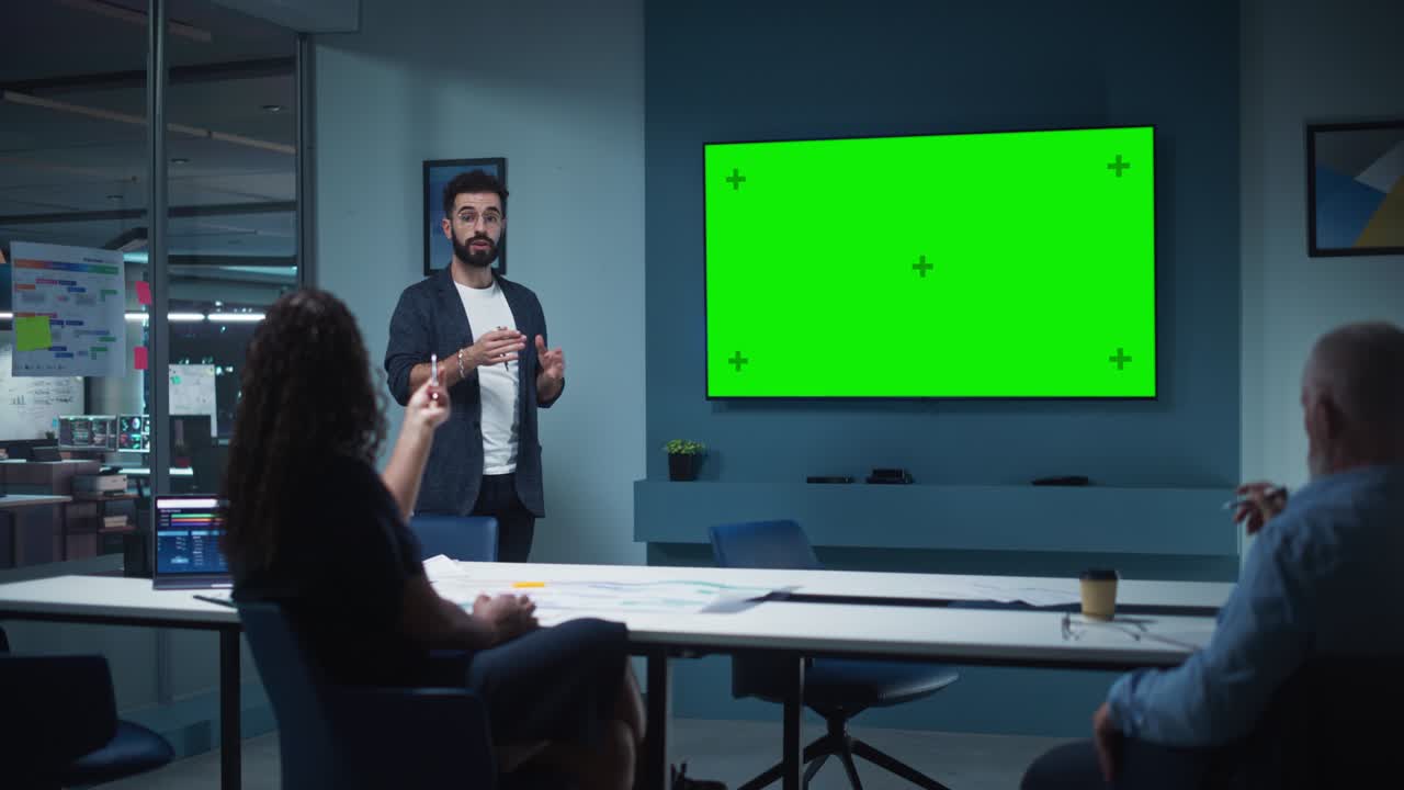 el gerente de operaciones de la compañía realiza una presentación de reunión para un equipo de ejecutivos. hombre adulto usa un televisor ancho con pantalla verde horizontal. la gente trabaja en una oficina de negocios.