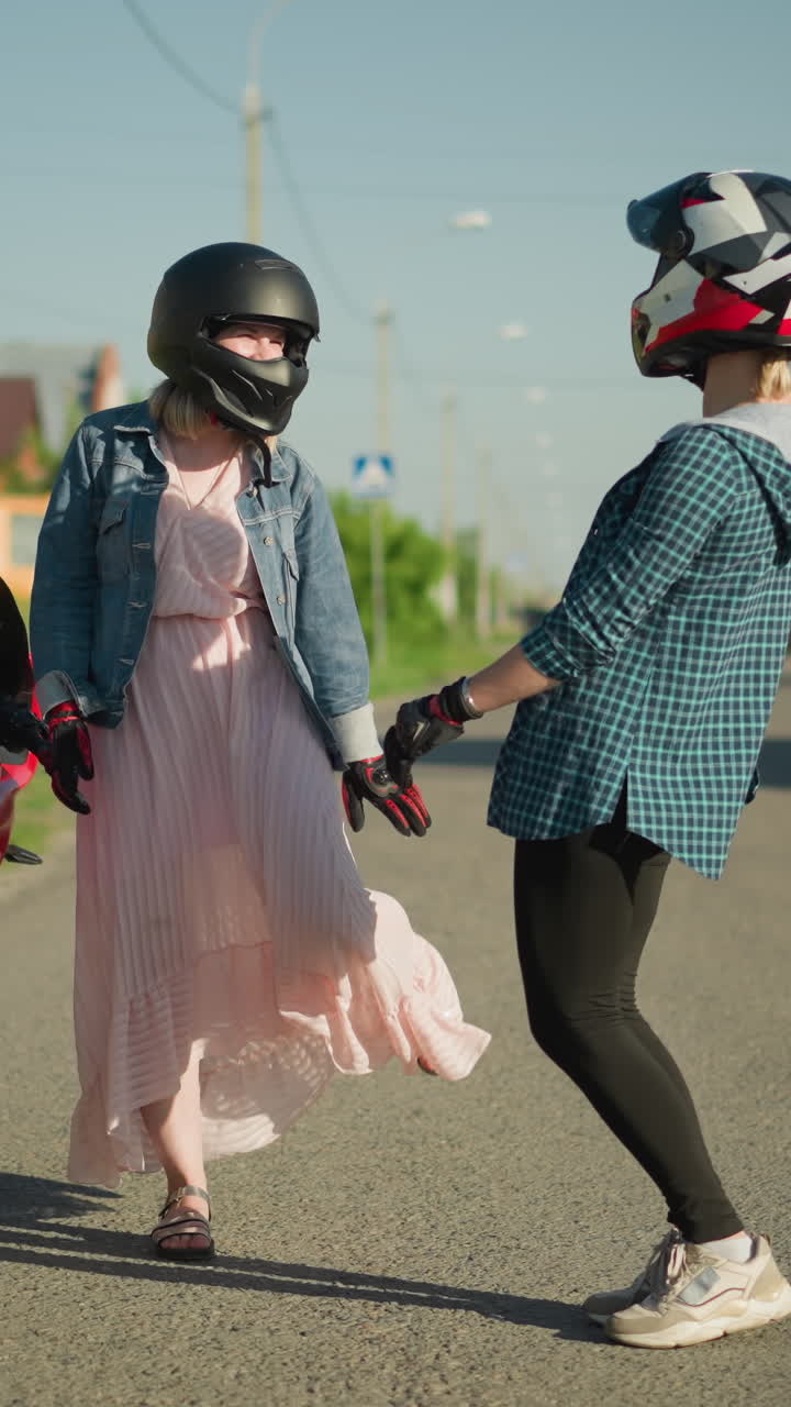 dos amigos con cascos de pie junto a una bicicleta eléctrica estacionada, compartiendo un momento alegre, una está jugando con su vestido rosa mientras la otra se ríe, postes eléctricos, casas y vegetación