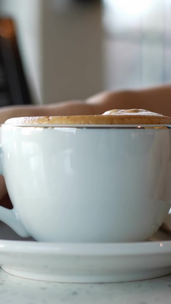 Video verticale: Mano destra che si allunga toccando tazza e piattino bianchi su tavolo maculato, sfiorando la crema