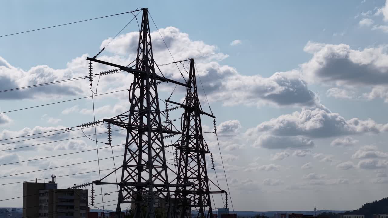 Two steel transmission towers and overhead power lines above city apartment blocks under dramatic clouds, showing electricity grid infrastructure and urban energy supply