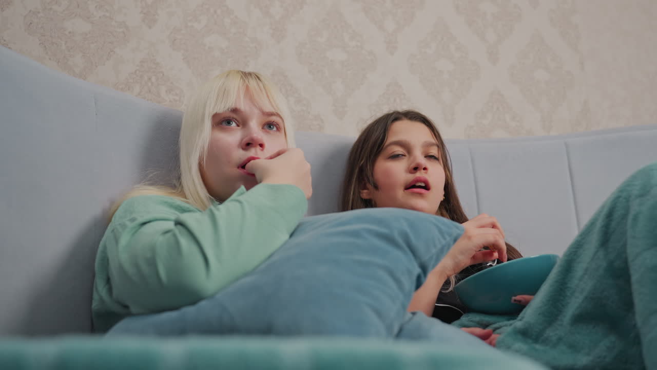 Two young women lie on bed closely wrapped in blankets, deeply focused on movie as one eats popcorn from bowl, creating scene of cozy indoor leisure and emotional engagement