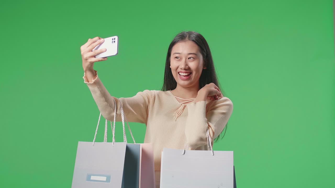 An Asian Shopping Woman With Shopping Bags Having Video Call On Mobile Phone While Standing In Front Of Green Screen