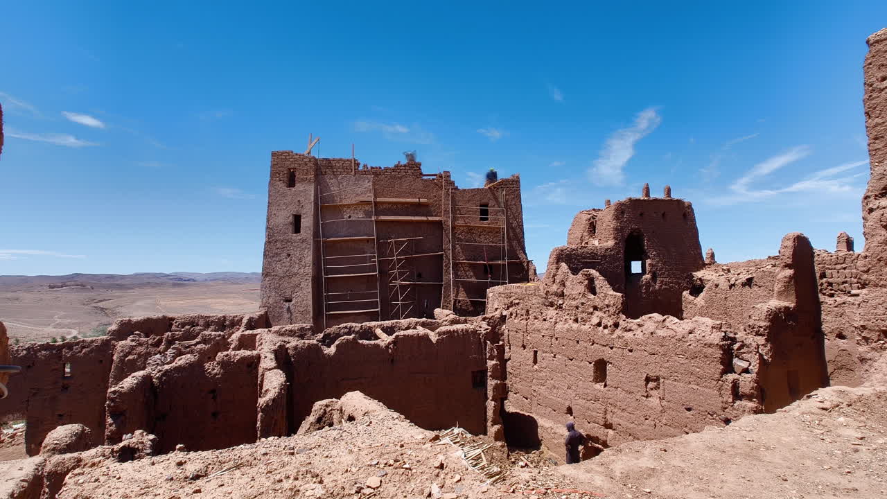 Traditional mud-brick architecture of a Moroccan ksar. Some structures are undergoing restoration with wooden scaffolding. The surrounding landscape is arid and hilly