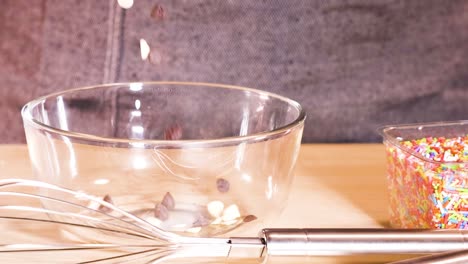 chocolate chips and colorful sprinkles are added to a glass bowl on a wooden surface.