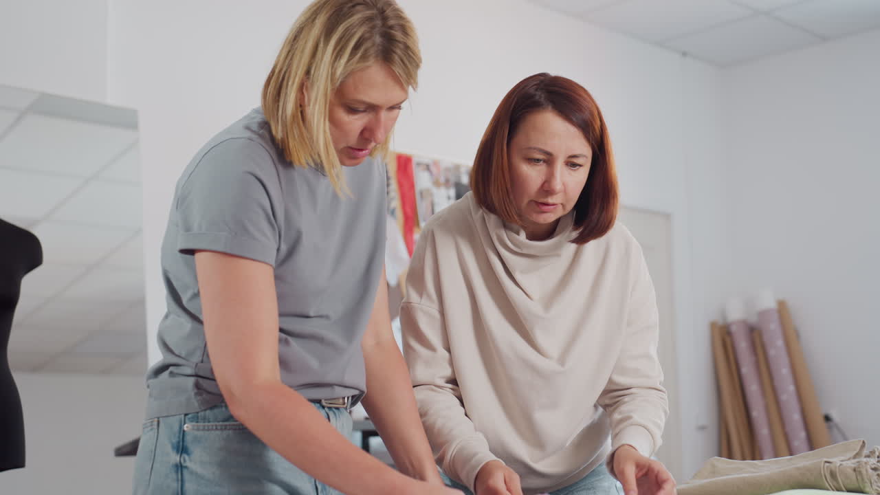 Fashion designers matching different fabric colors while standing at work table in bright creative workspace surrounded by colorful textiles fabric rolls mannequin sewing tools and design sketches