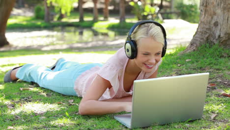 a woman lies in the park with headphones on while listening to a laptop