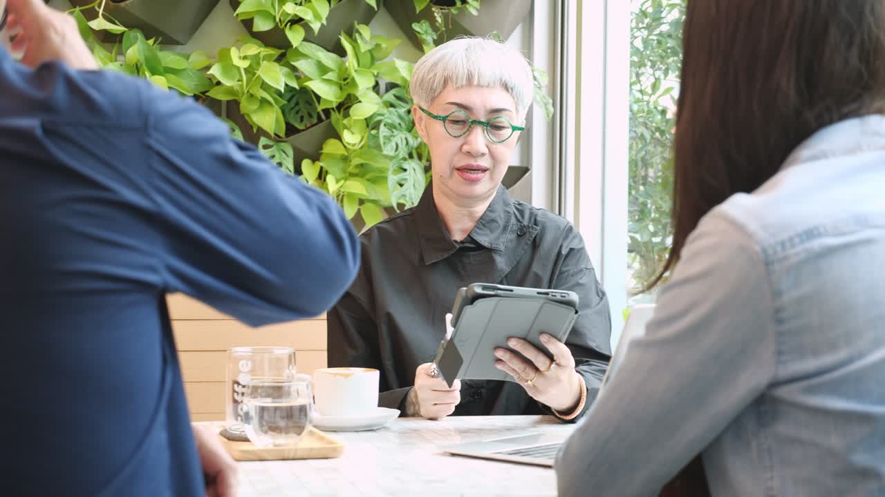 En ældre kvinde diskuterer en projektaftale og underskriver på en tablet med forretningsfolk på en café. Samtalen og aftalen om forretningspartnerskab foregår i kaffebaren, med en rådgiver og agent.