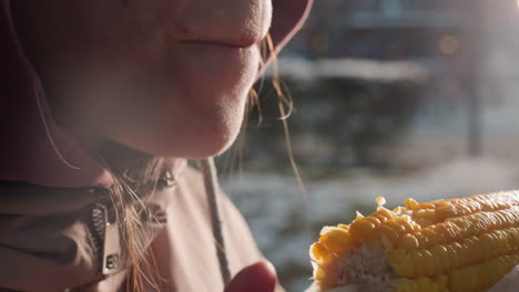 close-up of person eating corn held by stick wrapped in white plastic with bokeh lights in blurred background, steam rising from fresh corn in outdoor setting