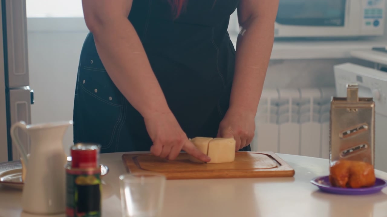 Mani adulte affettano formaggio rotondo su un tagliere di legno in una luminosa cucina di casa, poi dispongono i pezzi separati tenendo fermo il cibo, con grattugia, coltello, stoviglie e ingredienti freschi nelle vicinanze.