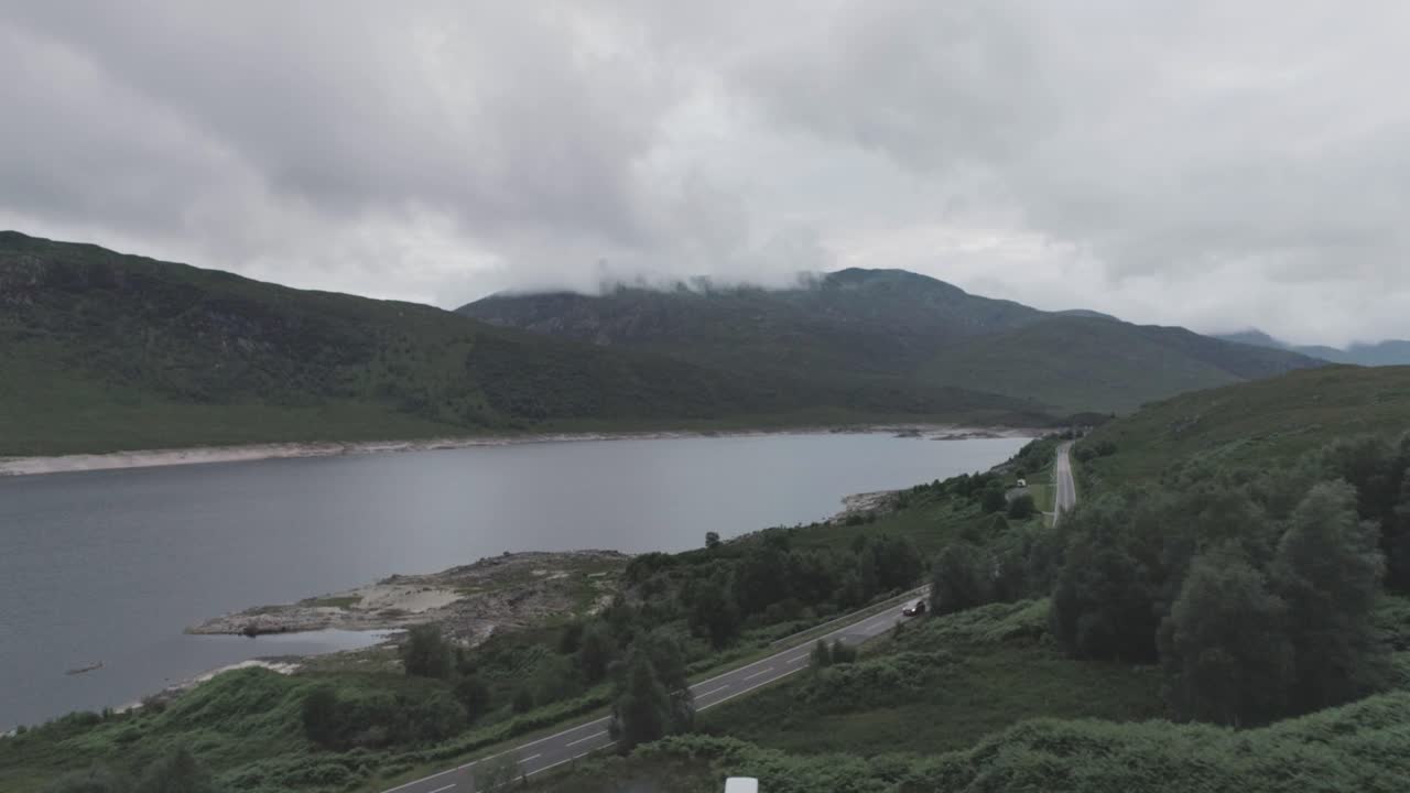 Wide drone shot showing Loch Beag and a campervan parked up for the night and a BBQ on the Loch edge with cloudy mountains in the background