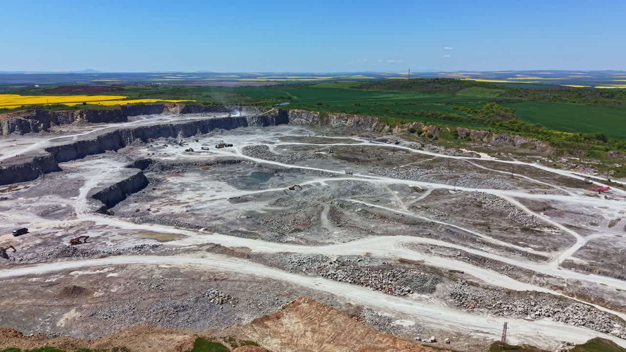 Cantiere minerario attivo a cielo aperto con macchinari pesanti al lavoro nella zona vicino a campi e colline sotto un cielo azzurro e sereno.