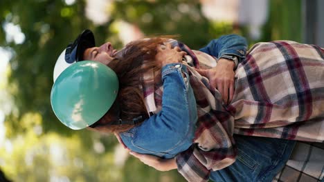 Vertical-video-of-a-happy-brunette-guy-with-curly-hair-in-a-moped-helmet-hugs-his-brunette-girlfriend-in-a-checkered-shirt-in-a-beautiful-summer-city