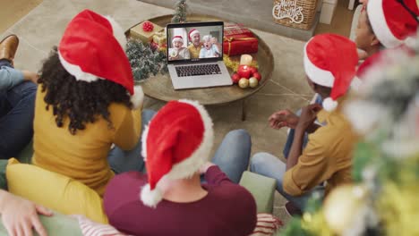 familia diversa con sombreros de santa usando una computadora portátil para una videollamada de navidad con familia sonriente en la pantalla