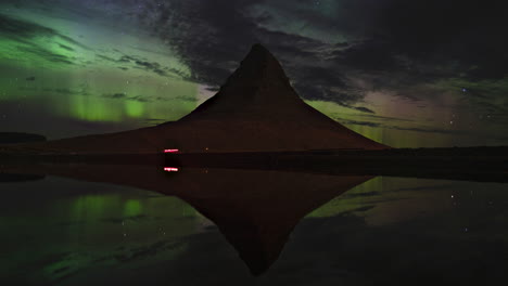 islandia, kirkjufell: lapso de tiempo de la aurora boreal con el reflejo del lago y los autos que pasan