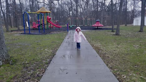 Una-Niña-Camina-Hacia-La-Cámara-Con-Un-Patio-De-Recreo-Al-Fondo-Por-Una-Acera-De-Hormigón-Bajo-La-Lluvia-Con-Un-Abrigo-Hinchado