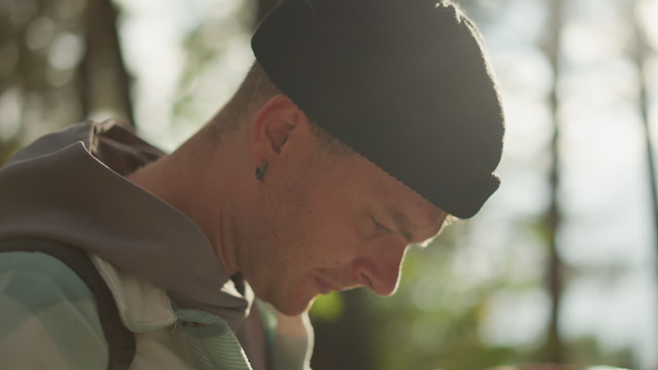 white guide focused on paper map, sunlit portrait among tall pines, concentrated expression, earring glint, hoodie and backpack straps visible, careful waypoint scanning, cinematic backlight creating