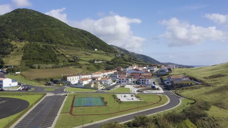 Velas,-Sao-Jorge-island,-Azores,-Portugal-view-of-village-with-whitewashed-houses,-skate-park-and-sports-field,-with-surrounding-mountains-and-vegetation-orbital-High-quality-4k-drone-footage