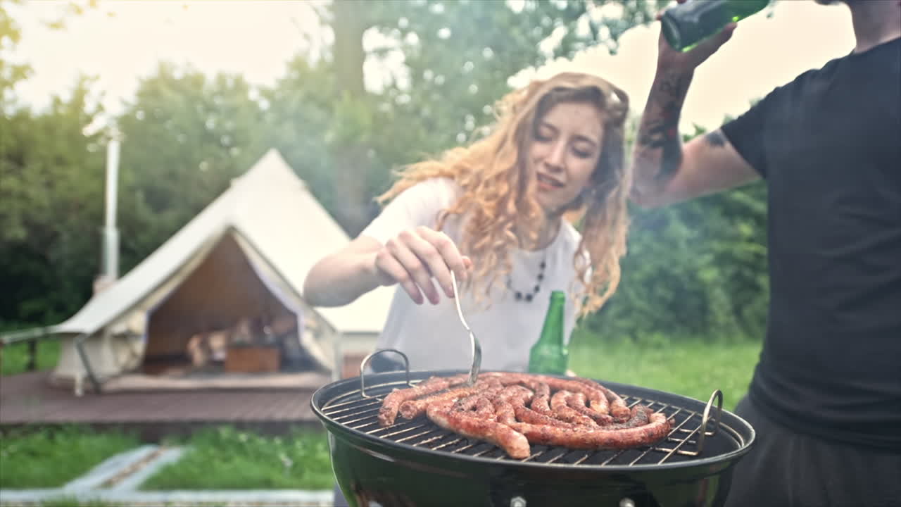 Giovane coppia che frigge carne alla griglia e beve birra. Tanto verde intorno. Glamping. Slow motion